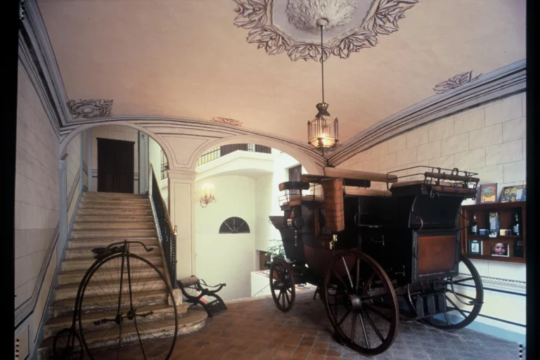 Interior del Museu Romàntic de Sitges con un carruaje histórico, una bicicleta antigua y una escalera noble con techo decorado.