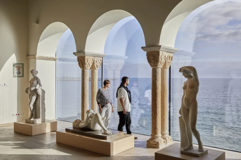 Dos visitantes contemplan esculturas novecentistas de Joan Rebull en la sala del Mirador del Museo de Maricel de Sitges, con el Mediterráneo de fondo.
